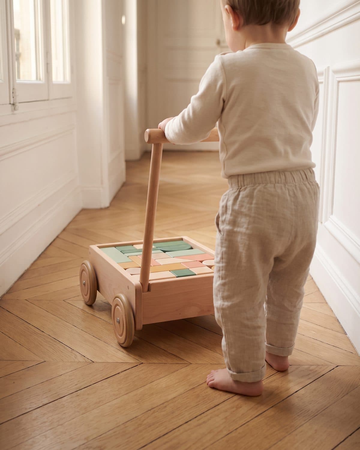 Un tout-petit, vu de dos, pousse le chariot Petit Pas dans le couloir parqueté d'un appartement